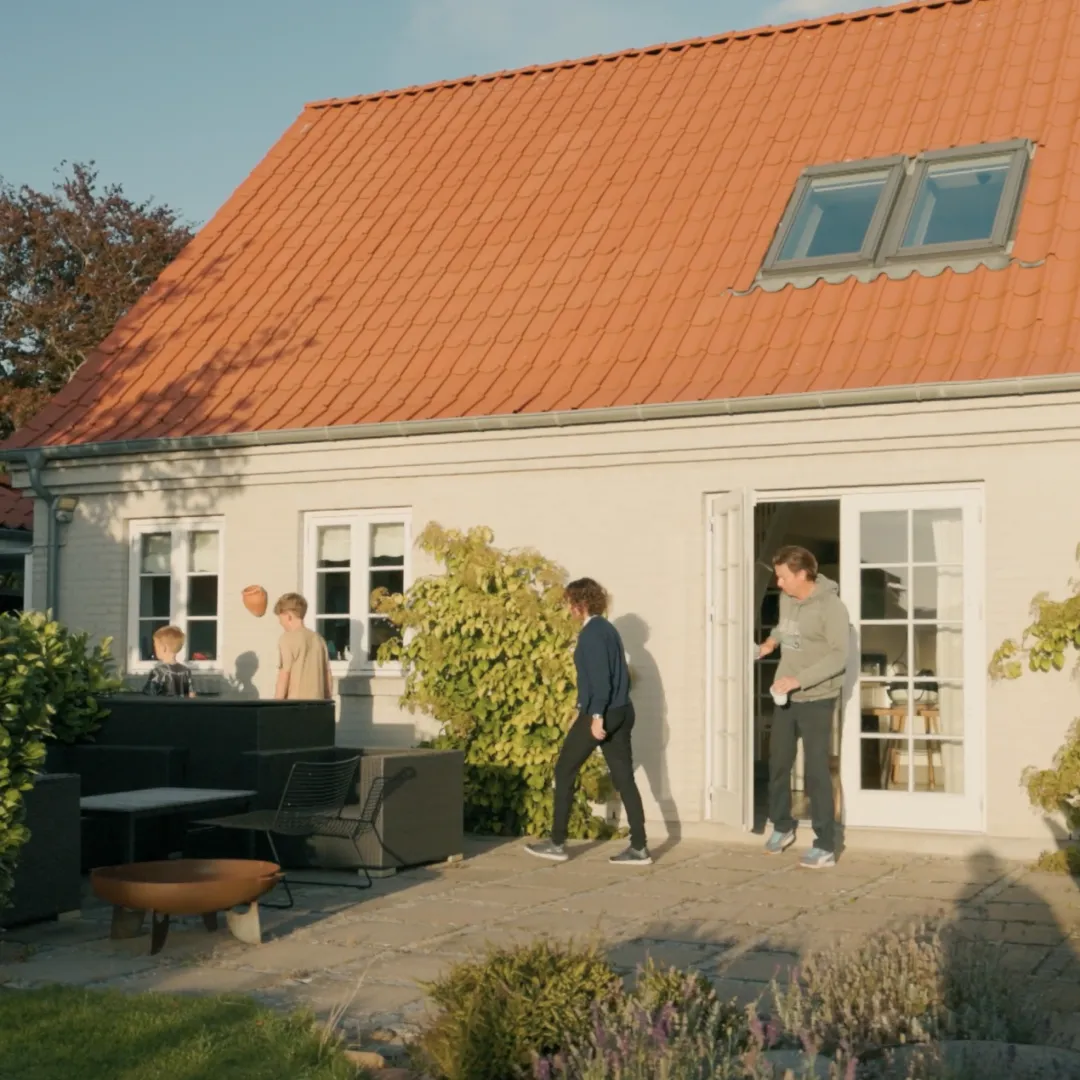 Four people on a sunny patio outside a small house with a red roof, open doors, and greenery.