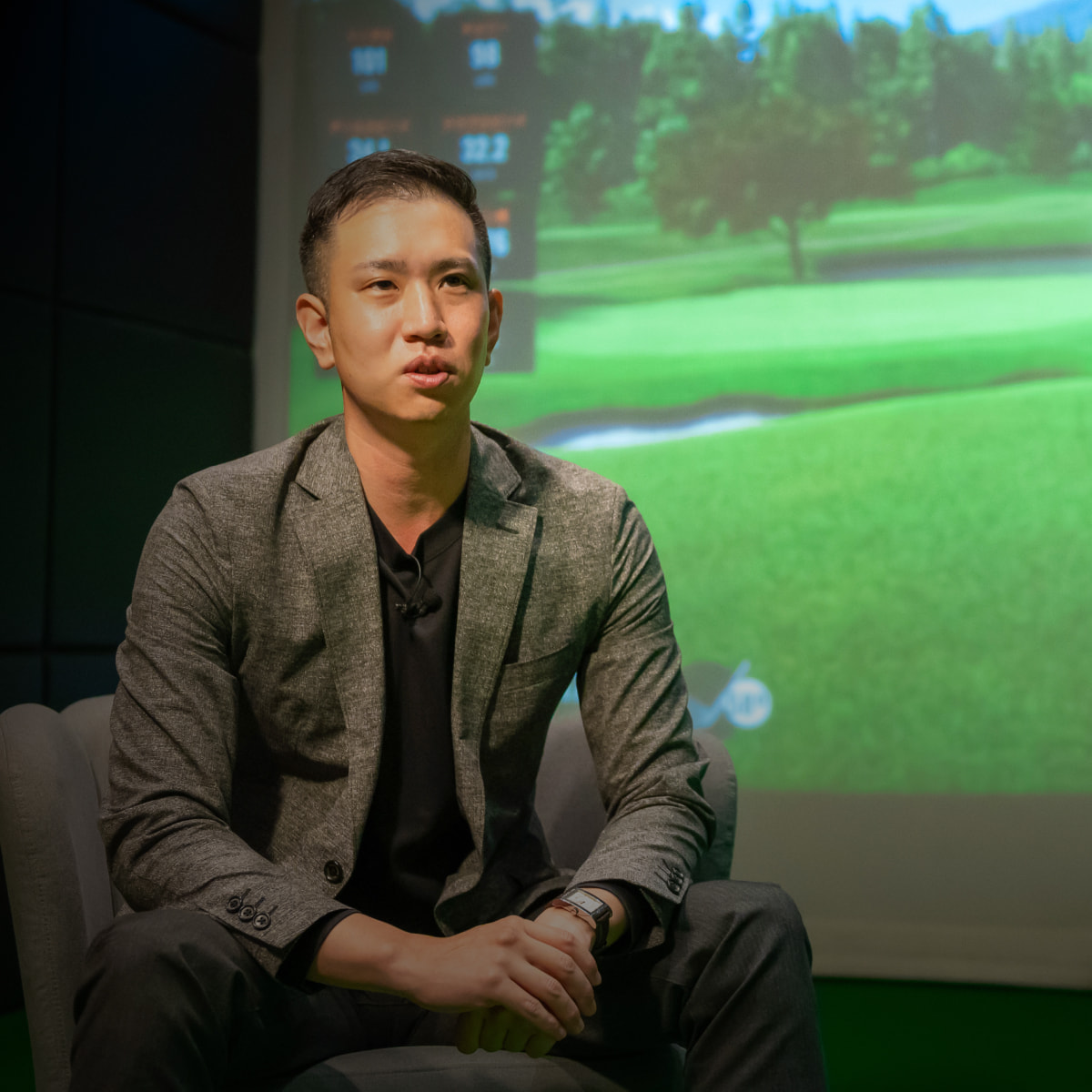 Person in a gray suit sitting on a chair, speaking, with a golf course image projected in the background.