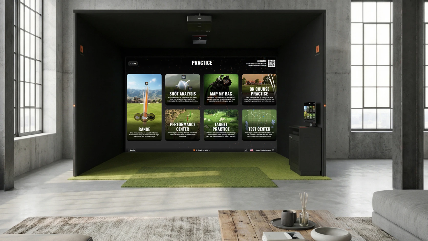 Indoor golf simulator setup with a large screen displaying practice options and lush green turf, set in a modern room with concrete walls.