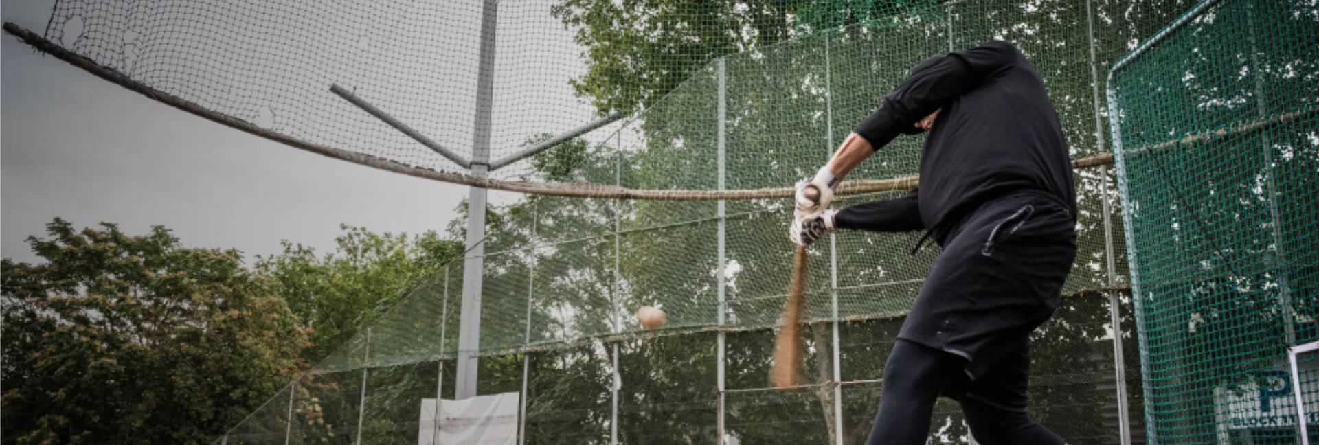 Person in black sportswear swings a baseball bat at a ball in a fenced outdoor area, surrounded by trees and netting.