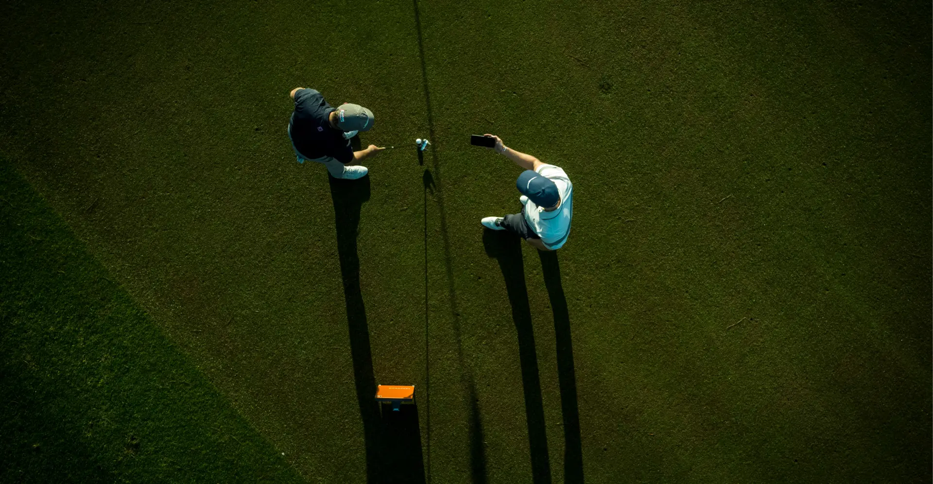 Aerial view of two golfers on a green course, one taking a selfie. Long shadows cast across the grass in the sunlight.