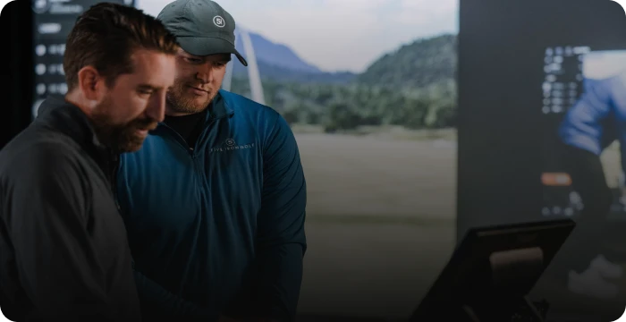 Two men in casual outdoor attire look at a computer screen, with a landscape image displayed in the background.