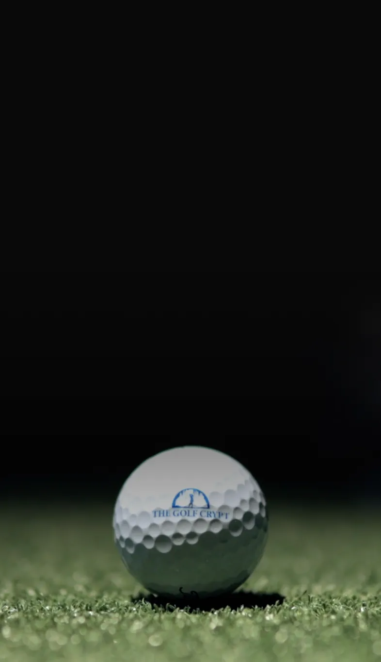 Close-up of a white golf ball displaying "The Golf Crypt" logo, sitting on green turf with a dark, out-of-focus background.