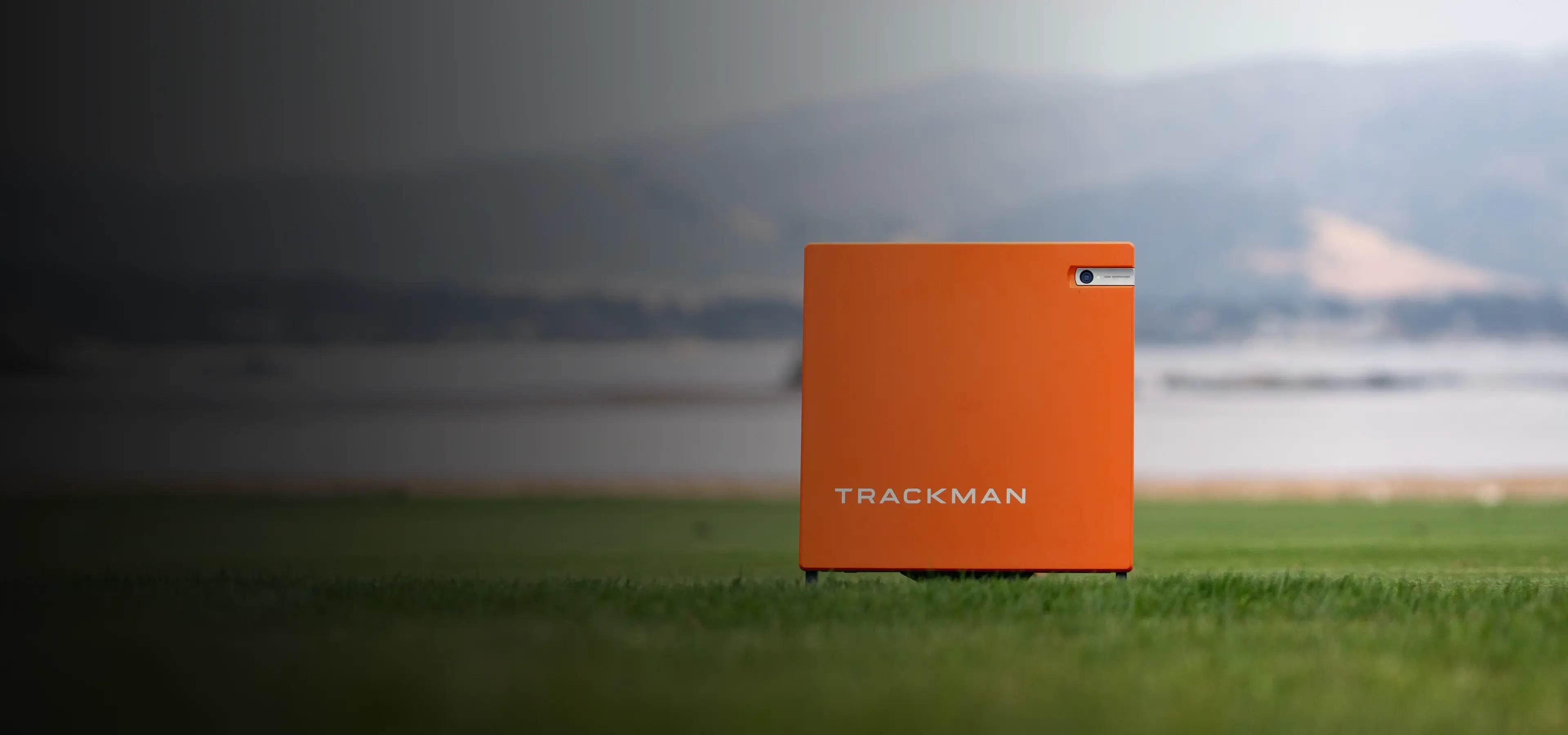 An orange TrackMan device on grass with a blurred mountainous landscape in the background.