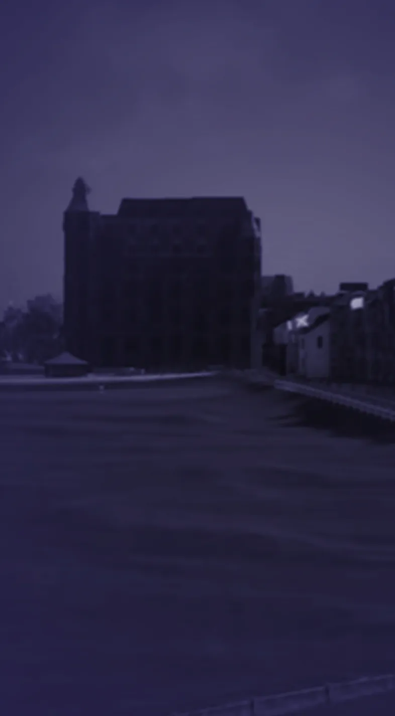 A dark, silhouetted building and houses under a purple night sky, with a calm body of water in the foreground.